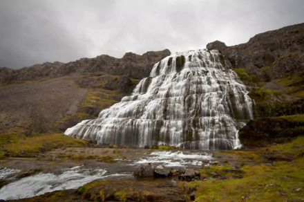 Dynjandi waterfall in Iceland cascades down rugged cliffs under a cloudy sky, set amidst green and rocky terrain, captured in a high-definition desktop wallpaper.