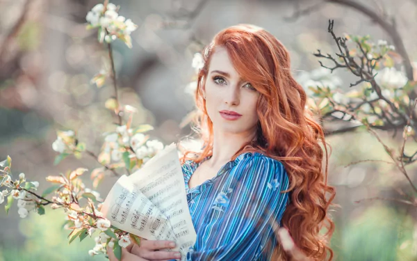 Redheaded woman with green eyes holds sheet music outdoors among blooming branches, captured in a soft bokeh background for an HD desktop wallpaper.