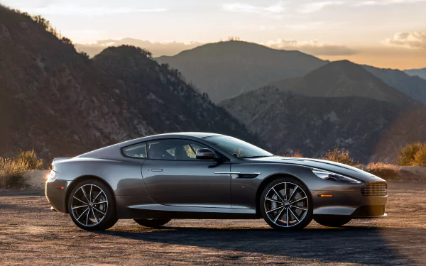 Silver Aston Martin DB9 grand tourer vehicle on a mountain overlook at sunset, 4K Ultra HD PC desktop wallpaper.