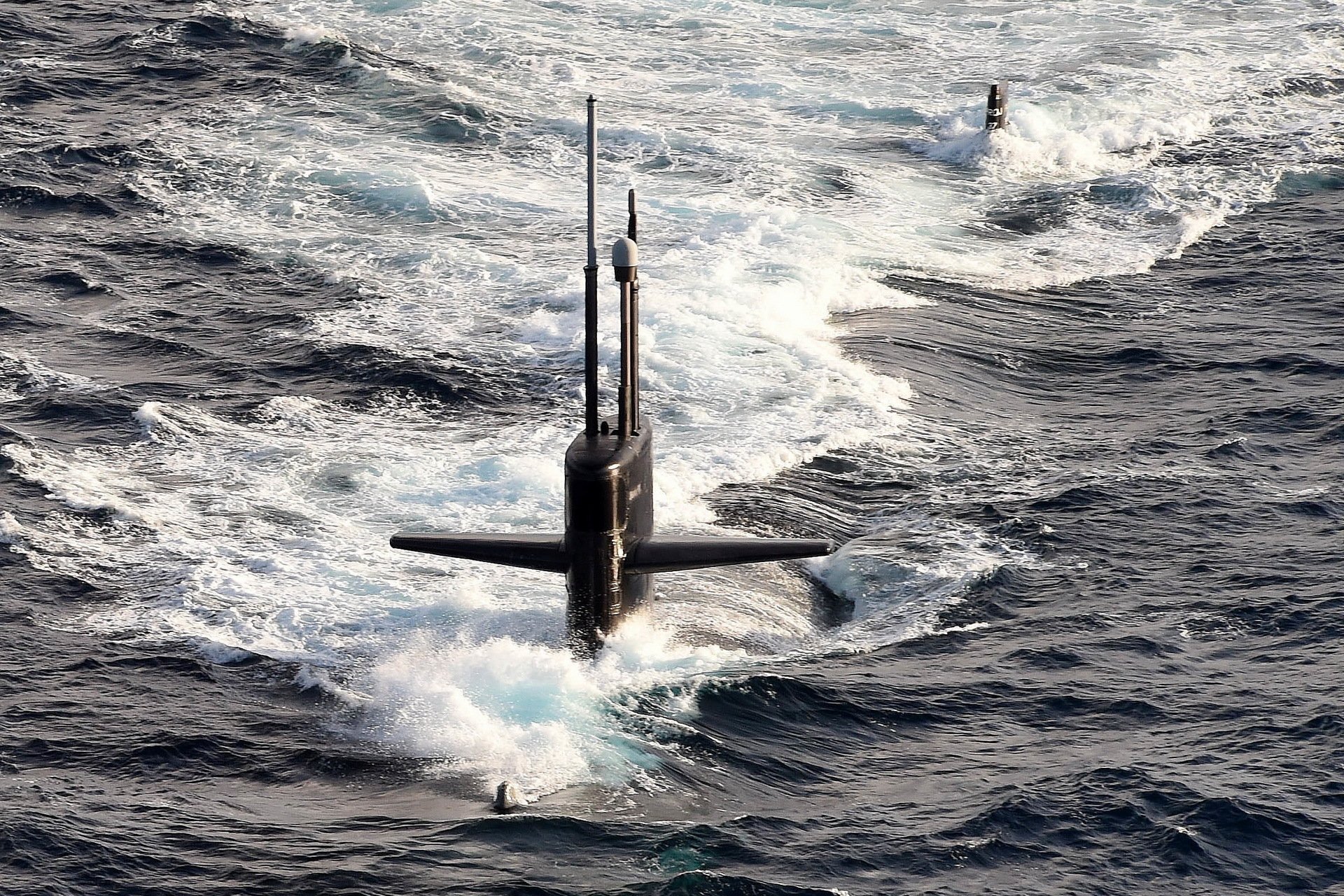 HD desktop wallpaper of a military submarine surfacing through ocean waves, capturing the vessel's conning tower and antennas in dynamic motion.
