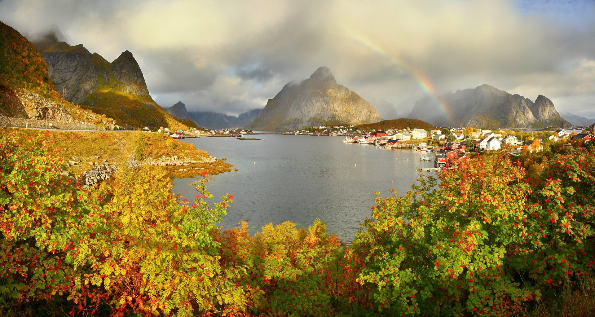 HD desktop wallpaper featuring the village of Reine in Lofoten, Norway, with mountains, a lake, and a vibrant rainbow arching across the cloudy sky.