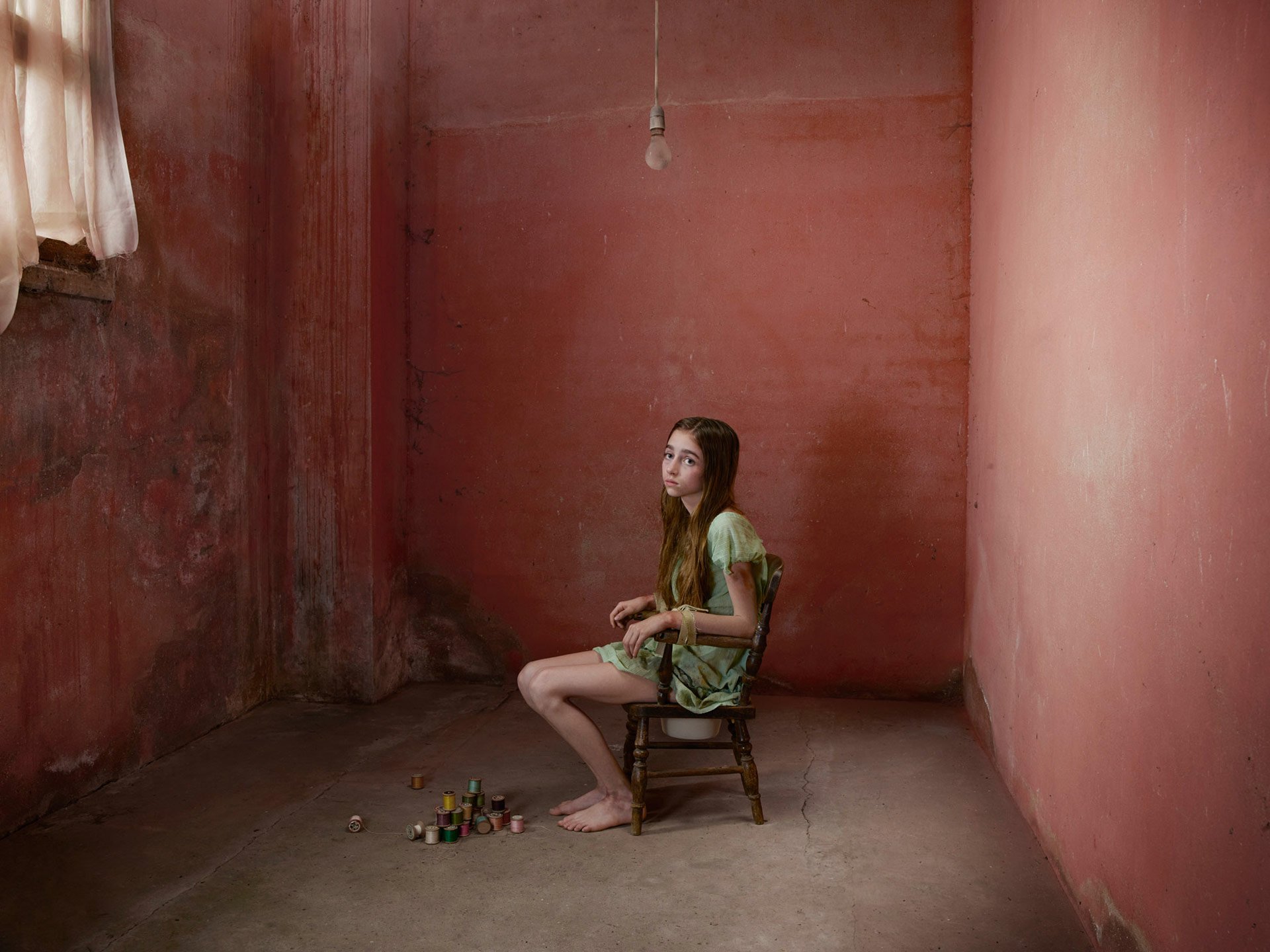 A young girl in a green dress sits on a wooden chair in a dim, empty room with pink walls, creating a quiet, fantasy-inspired fairy tale atmosphere in this HD desktop wallpaper.