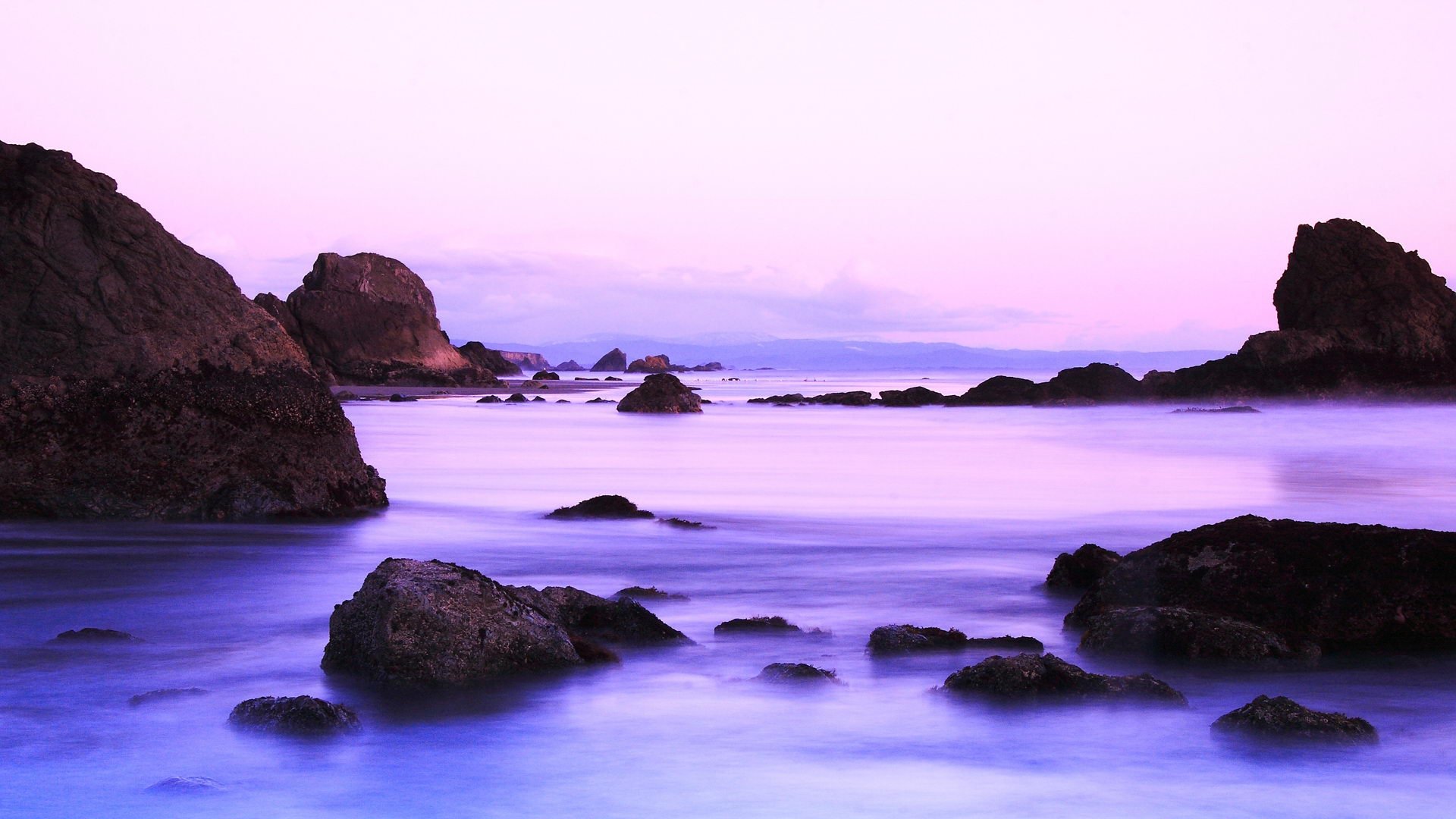 A serene beach scene featuring tranquil waters and gentle waves, surrounded by rocky formations under a pastel sky. This HD wallpaper captures the beauty of nature.