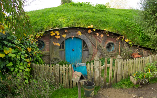 HD desktop wallpaper of a man-made Hobbiton house in Comarca, featuring a round blue door set into a grassy hillside with rustic wooden fencing and autumn decorations.