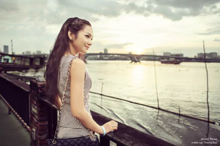 An Asian woman stands by a riverside railing, gazing at the sunset, with a cityscape in the background. This HD desktop wallpaper captures a serene and contemplative moment.