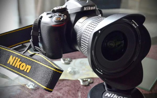 HD desktop wallpaper featuring a Nikon camera lens and body on a table, with a Nikon strap prominently displayed.