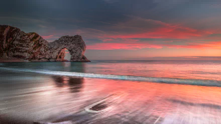 Sunset over the sea at Durdle Door, England, with the ocean gently washing onto the beach beneath a colorful sky on the horizon.