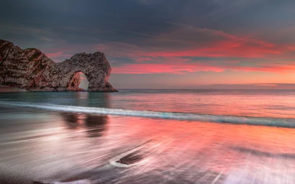 Sunset over the sea at Durdle Door, England, with the ocean gently washing onto the beach beneath a colorful sky on the horizon.
