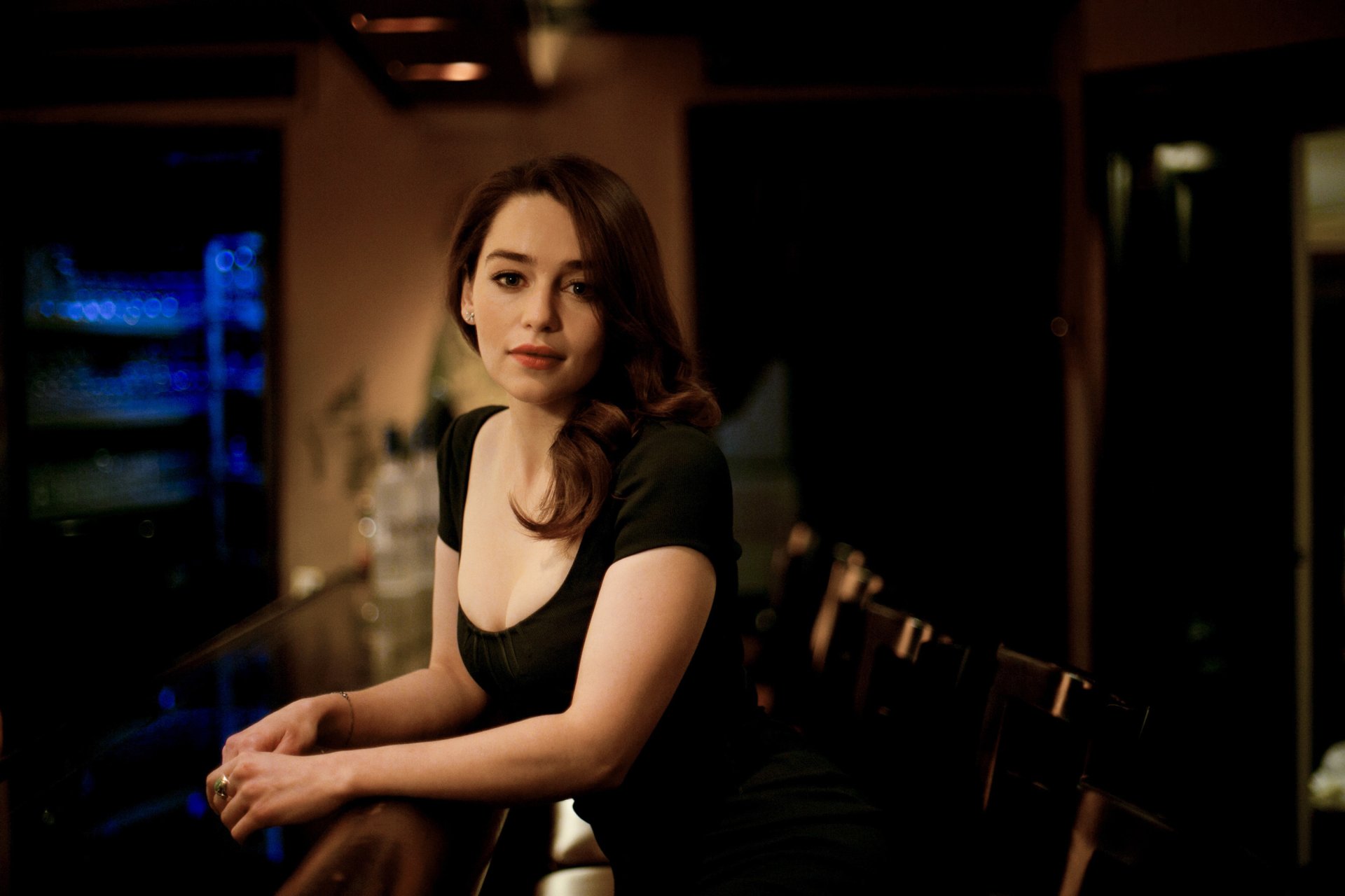 HD PC desktop wallpaper featuring celebrity Emilia Clarke seated indoors with a calm, poised expression against a softly lit background.