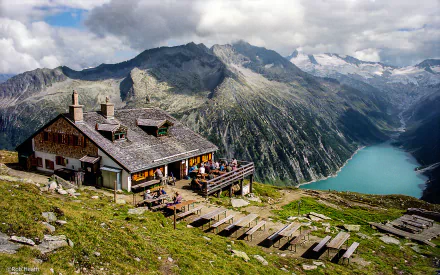 Mountain restaurant in Tyrol, Austria, perched on a grassy slope overlooking a turquoise alpine lake and rugged peaks under a partly cloudy sky.