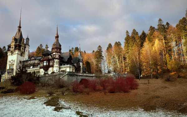 A stunning HD wallpaper of Peles Castle, nestled among autumn trees, with a mix of snow and earth, showcasing the beauty of man-made architecture in a natural setting.