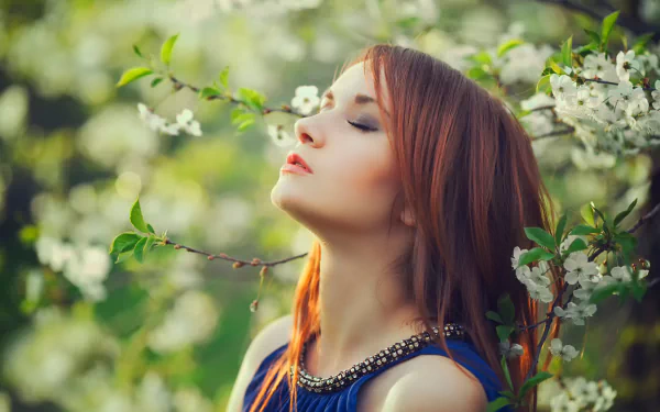 A redheaded woman in a blue top stands with eyes closed among blossoming white flowers on a sunny spring day, creating a serene mood. HD desktop wallpaper and background.