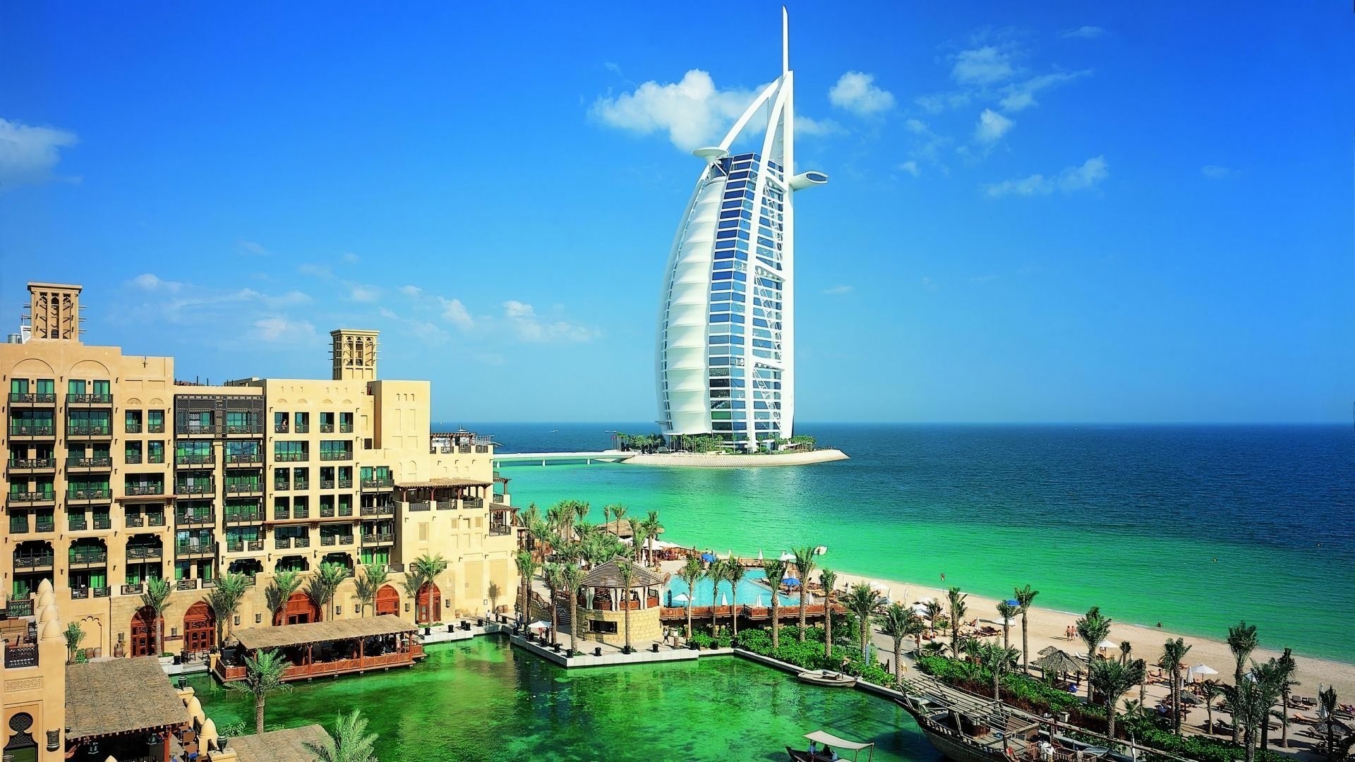 HD desktop wallpaper featuring Dubai’s iconic Burj Al Arab hotel rising over the ocean, surrounded by modern man-made architecture and vibrant coastal scenery.
