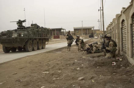 A military IAV Stryker armored vehicle moves along a dusty road, while soldiers engage in tactical maneuvers near a series of buildings, capturing a dramatic military scene in high definition.