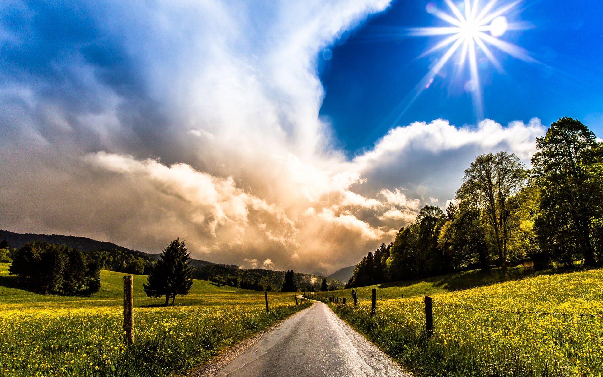 HD PC desktop wallpaper: country scene with a man-made road cutting through fields, trees along the roadside, dramatic clouds and bright blue sky with a sunburst.
