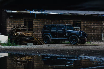 A black Jeep Wrangler SUV parked next to a rustic building, surrounded by stacked pallets and reflecting in a nearby puddle. A striking HD image for desktop wallpaper.