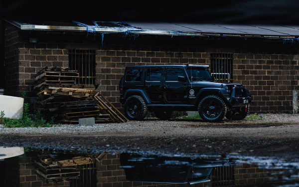 A black Jeep Wrangler SUV parked next to a rustic building, surrounded by stacked pallets and reflecting in a nearby puddle. A striking HD image for desktop wallpaper.