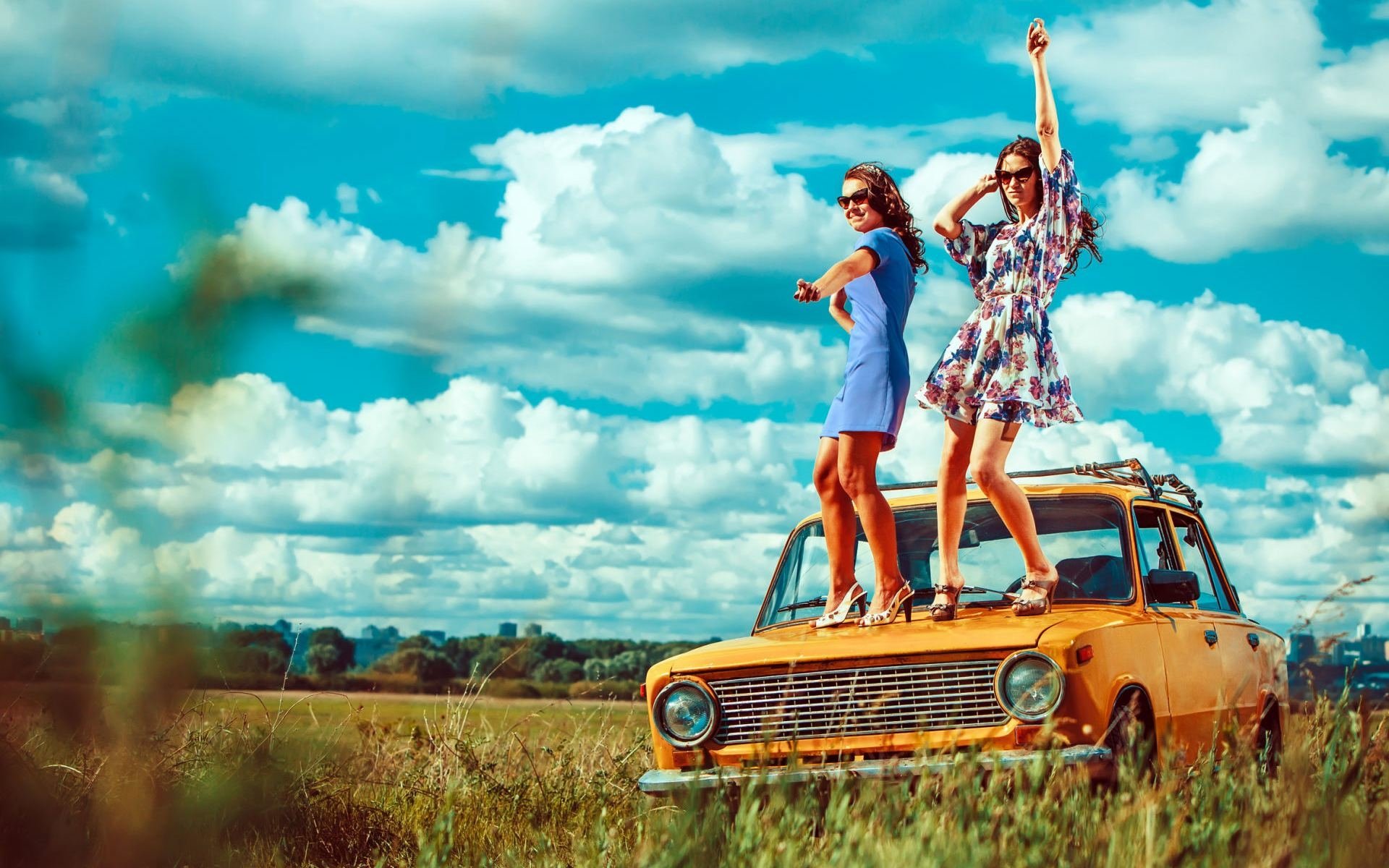 Two women in vibrant dresses stand joyfully on the roof of a vintage yellow car in a sunny field, captured in an HD desktop wallpaper radiating carefree mood.