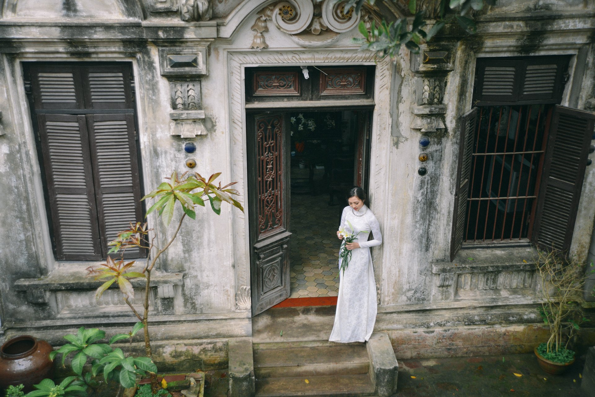 A Vietnamese woman in an elegant Ao Dai stands outside a rustic house, framed by greenery, capturing the essence of traditional culture in this vibrant 4K Ultra HD wallpaper.
