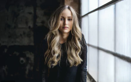 HD wallpaper featuring a beautiful blonde woman with long hair, standing in a dimly lit industrial setting with a large window.