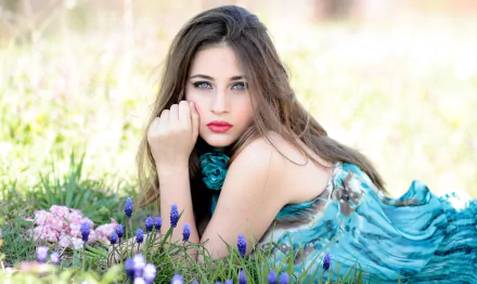 A brunette woman in a blue dress lies on the grass surrounded by purple flowers, gazing at the camera. The vibrant, serene scene creates a captivating HD desktop wallpaper and background.