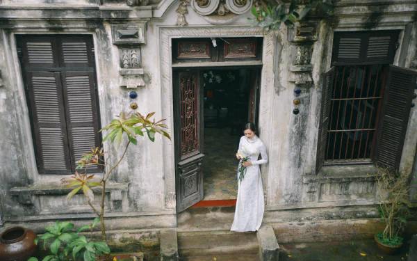 A Vietnamese woman in an elegant Ao Dai stands outside a rustic house, framed by greenery, capturing the essence of traditional culture in this vibrant 4K Ultra HD wallpaper.