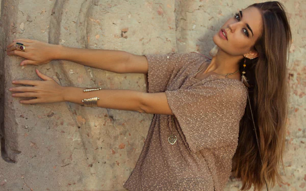 A Spanish woman named Clara Alonso poses elegantly against a textured wall, showcasing her long hair and stylish outfit in this 4K Ultra HD desktop wallpaper.