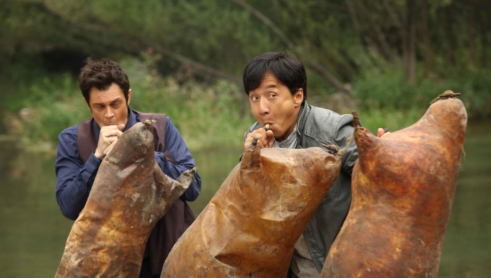 Jackie Chan and Renny Harlin in a comedic scene from the movie Skiptrace (2016), featuring the duo playfully interacting with large, oddly shaped objects.