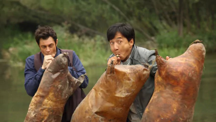 Jackie Chan and Renny Harlin in a comedic scene from the movie Skiptrace (2016), featuring the duo playfully interacting with large, oddly shaped objects.