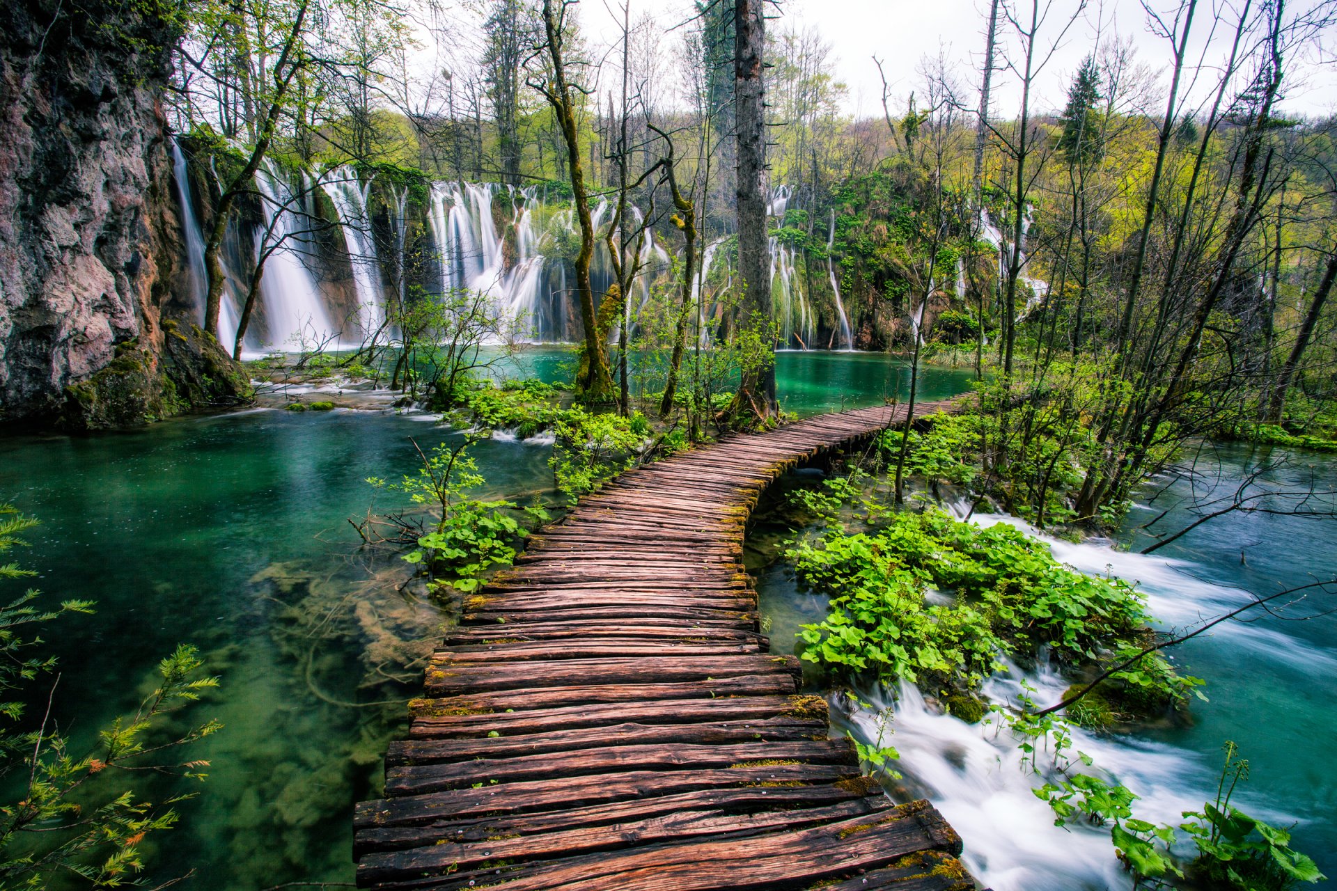 HD PC desktop wallpaper - wooden walkway bridge winding through trees and turquoise pools beside cascading waterfalls at Plitvice Lakes National Park, lush nature scene.