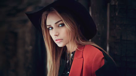 A woman with blue eyes and long blonde hair wears a black hat and an orange blazer, gazing intently. This HD image serves as a stunning desktop wallpaper or background.