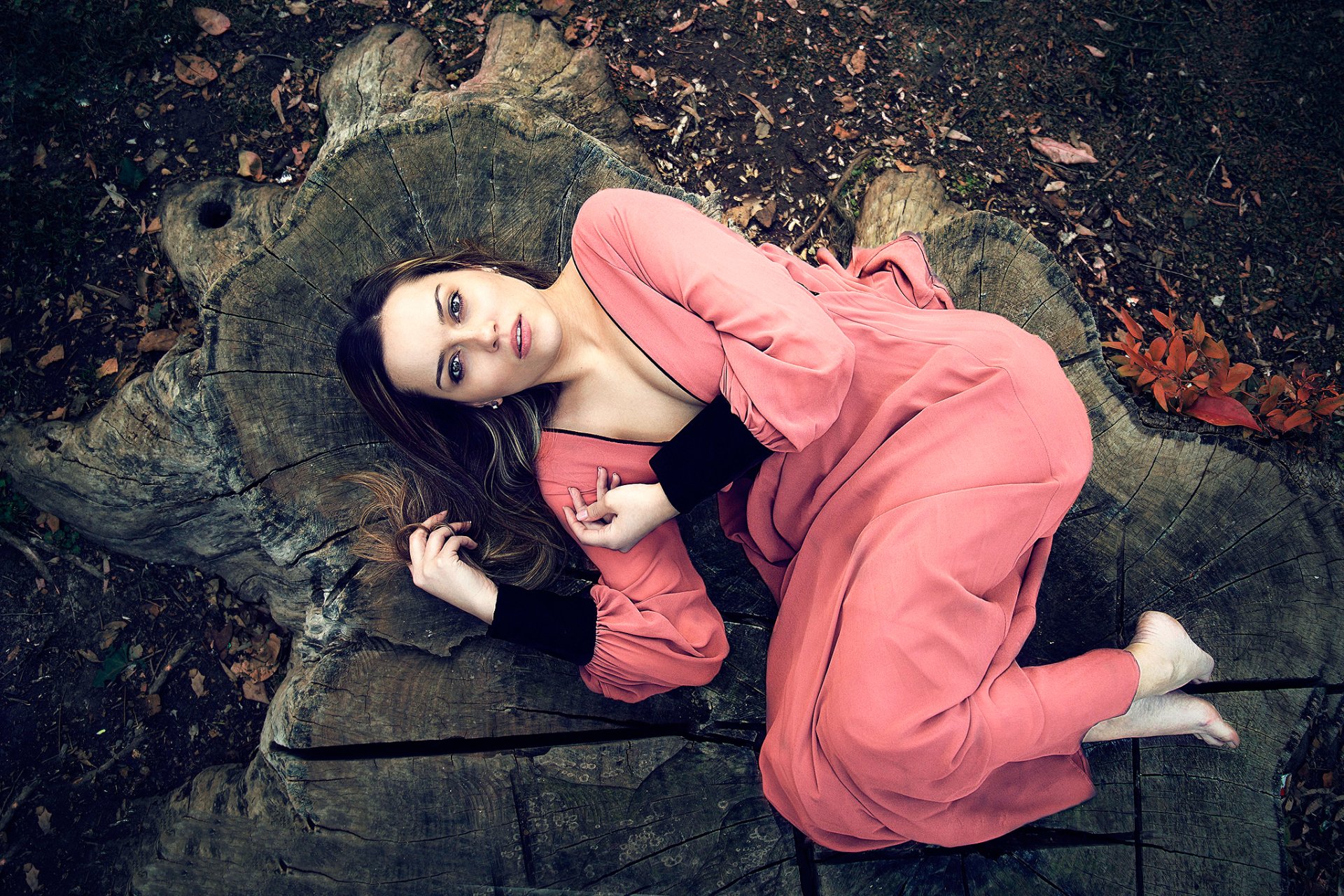 A brunette woman in a flowing pink dress lies thoughtfully on a large tree stump outdoors, creating a moody and serene HD desktop wallpaper background.