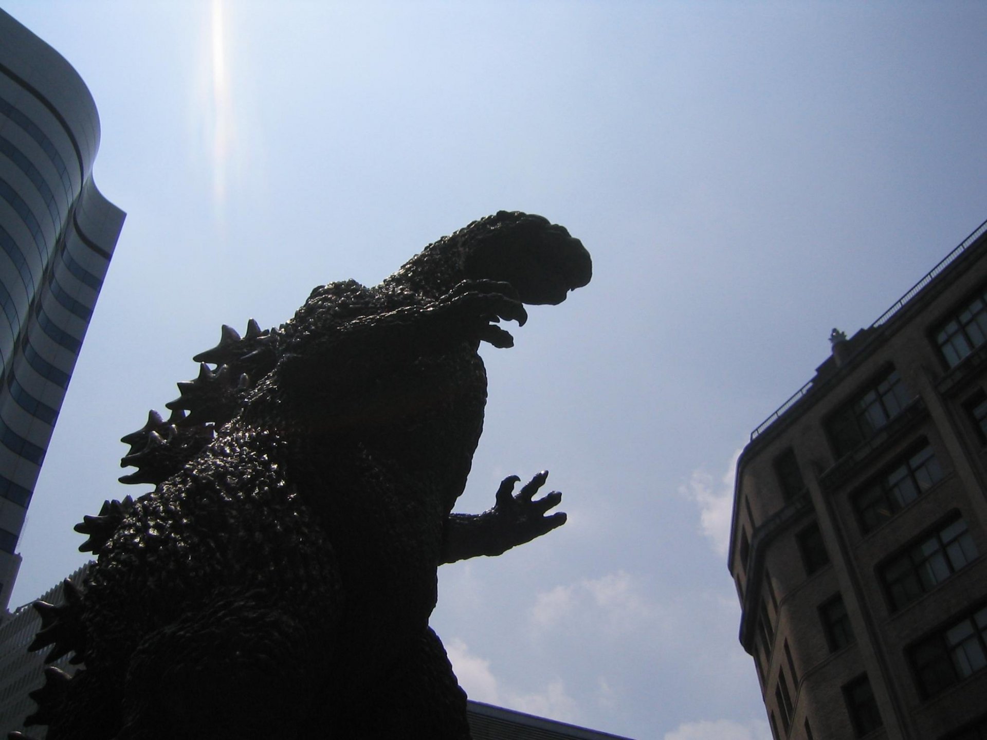 A striking HD desktop wallpaper featuring Godzilla in silhouette against a clear blue sky, poised among city buildings, capturing the iconic movie monster's formidable presence.