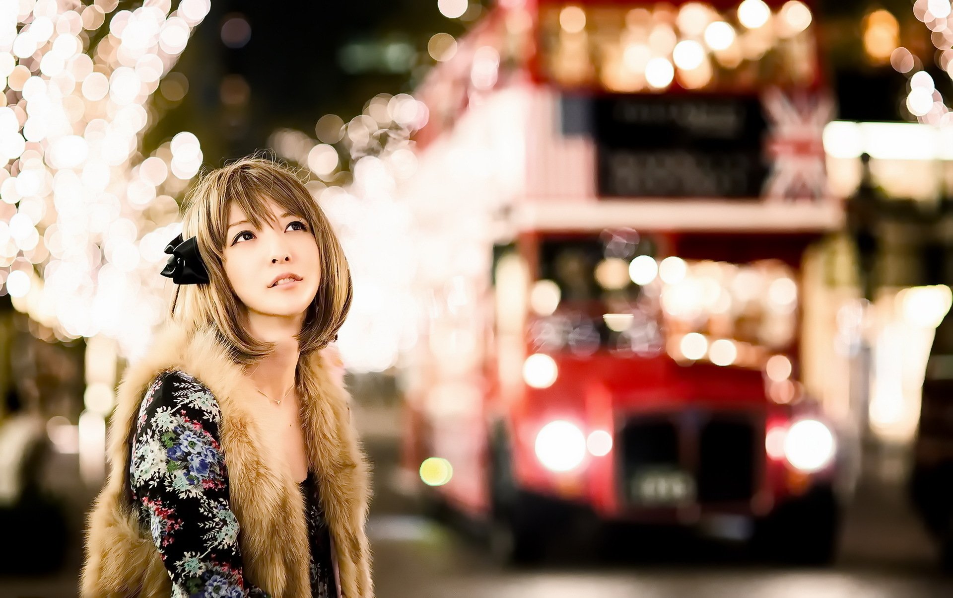 A high-definition wallpaper featuring an Asian brunette woman with brown eyes in a festive cityscape at night, standing in front of a red double-decker bus. The background is adorned with bokeh lights.