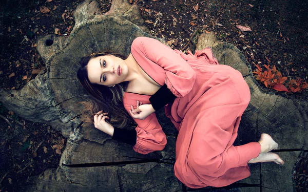 A brunette woman in a flowing pink dress lies thoughtfully on a large tree stump outdoors, creating a moody and serene HD desktop wallpaper background.