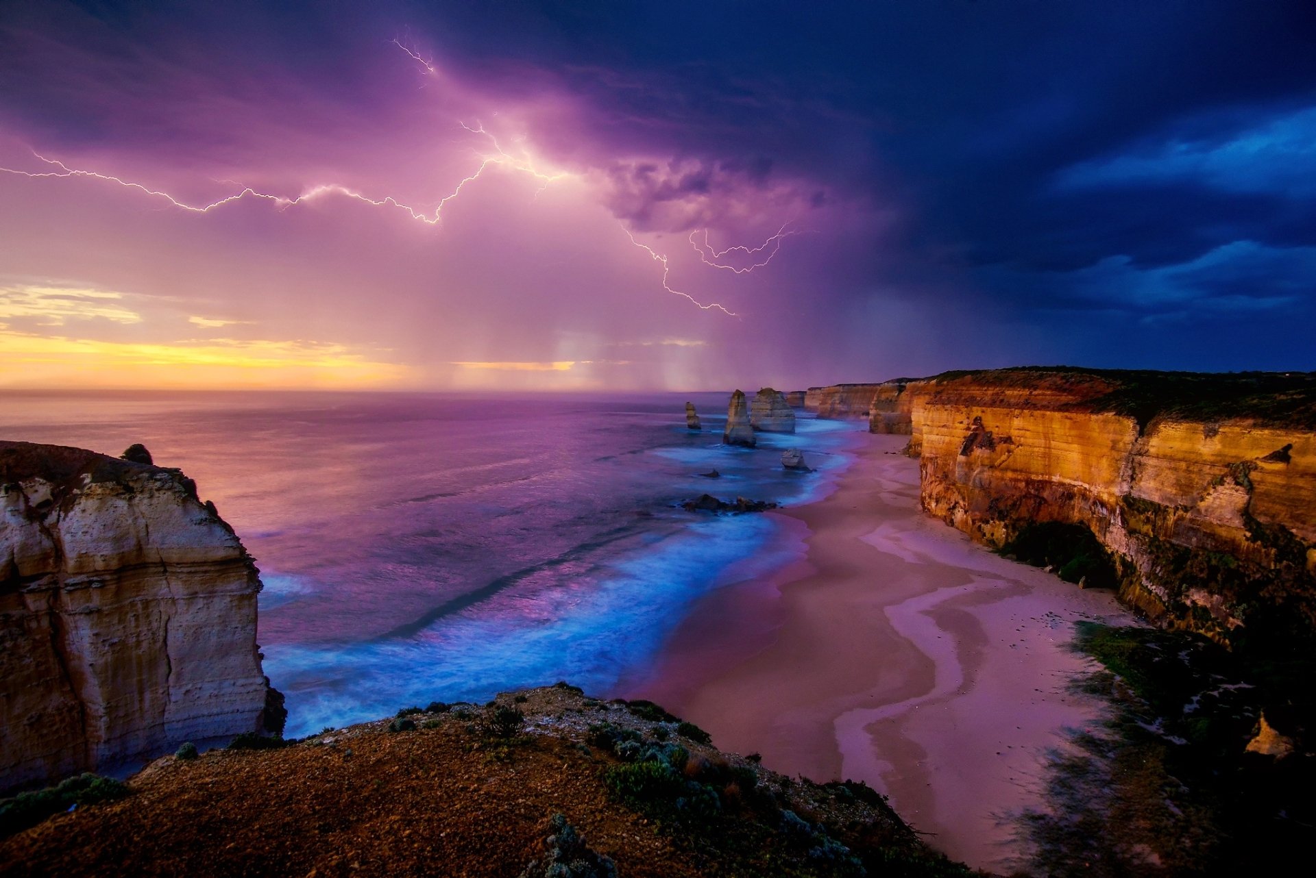 Dramatic lightning illuminates the sky over the sandy beach and sea at The Twelve Apostles, Australia, with the ocean horizon visible in this HD desktop wallpaper.