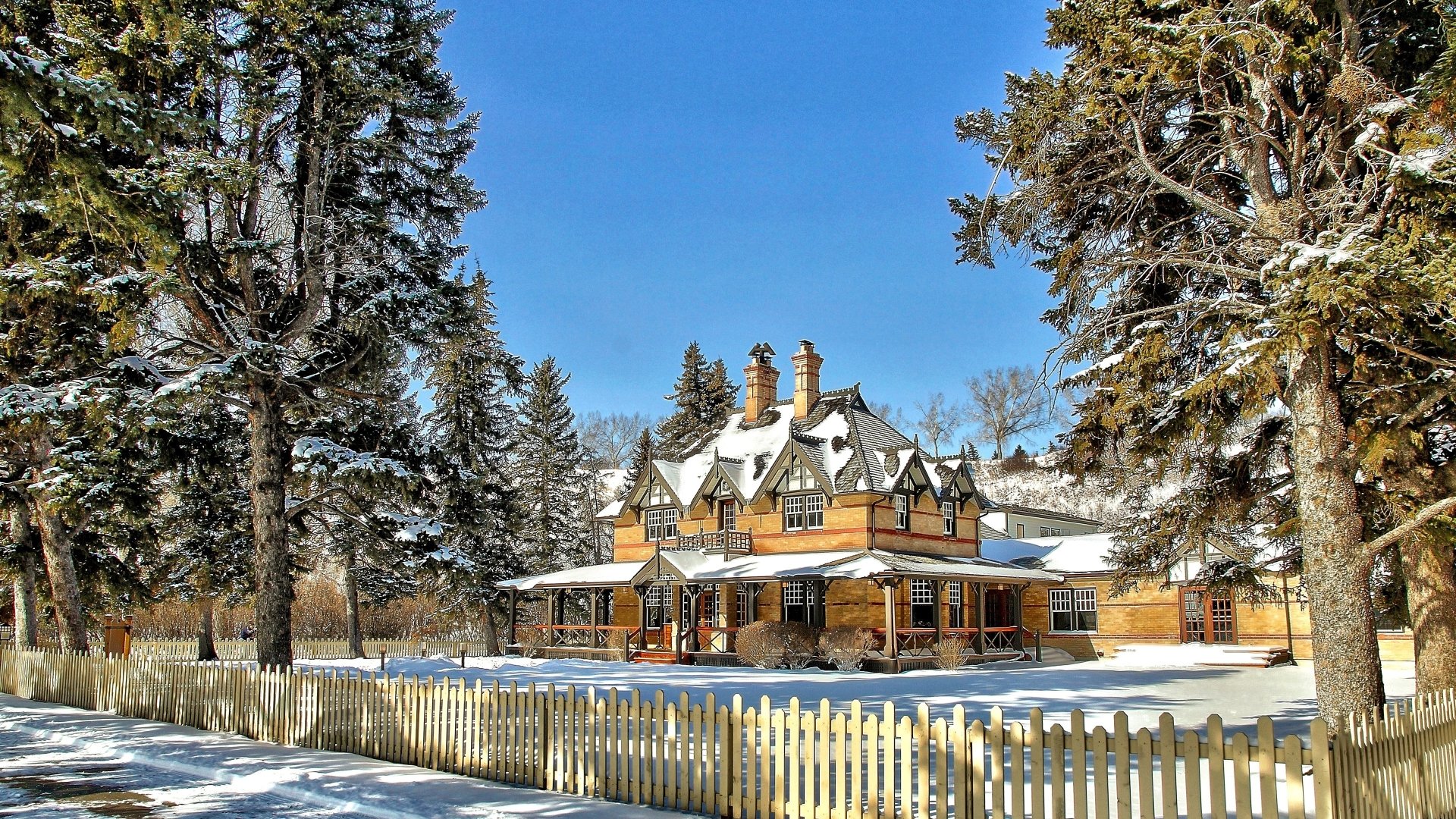 A 4K Ultra HD desktop wallpaper featuring a snow-covered mansion surrounded by trees and a wooden fence under a clear blue sky.