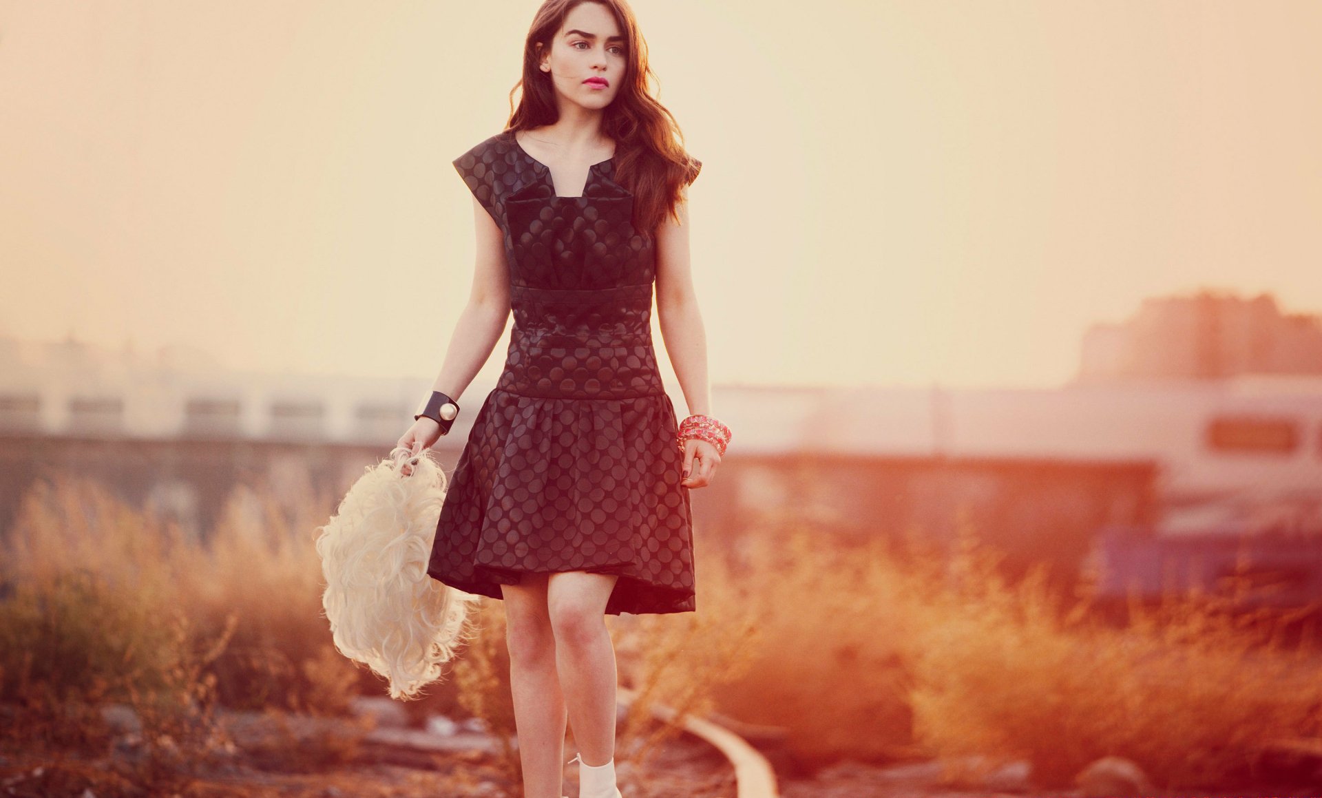 Brunette English actress Emilia Clarke in a black dress with lipstick, walking outdoors at sunset, featured in a HD PC desktop wallpaper and background.