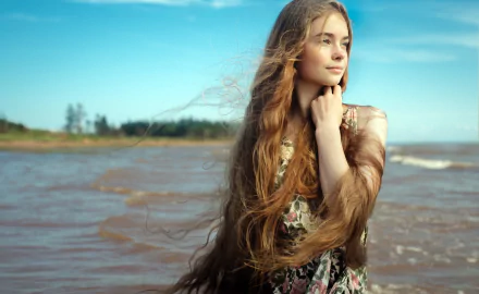 A woman with long blonde hair and blue eyes stands by the beach, her hair blowing in the wind. This HD desktop wallpaper captures her serene mood against a backdrop of sea and sky.