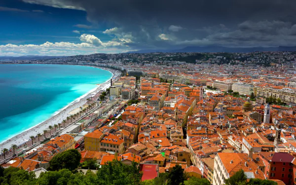 Panoramic HD desktop wallpaper of Nice, France: Mediterranean turquoise bay, pebbled beach and man-made seafront beside terracotta-roofed town.