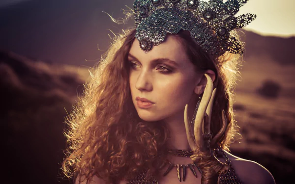 HD PC desktop wallpaper of a redheaded model with green eyes wearing an ornate crown and layered necklace, gazing across sunlit desert hills.