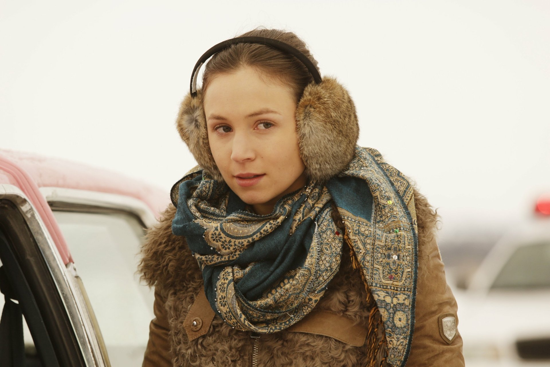 Wynonna Earp TV show 2K Quad HD desktop wallpaper — young woman in earmuffs and patterned scarf leans by a car in a snowy, muted landscape.