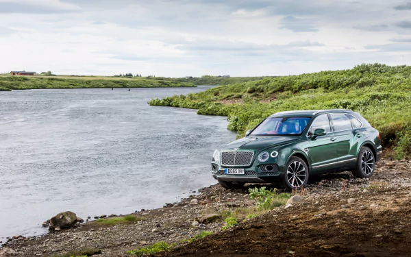 A green Bentley Bentayga SUV parked by a riverbank, surrounded by lush greenery, showcasing its sleek design in a serene landscape. 4K Ultra HD quality wallpaper.