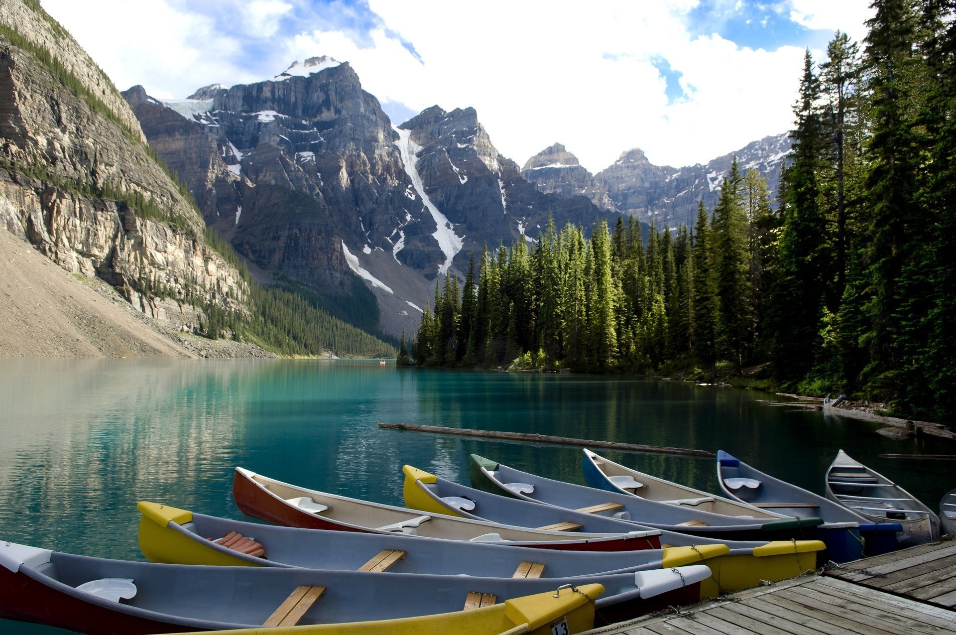 HD desktop wallpaper of colorful canoes docked at Moraine Lake, surrounded by pine trees and towering mountains under a partly cloudy sky.