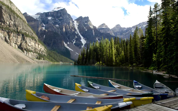 HD desktop wallpaper of colorful canoes docked at Moraine Lake, surrounded by pine trees and towering mountains under a partly cloudy sky.
