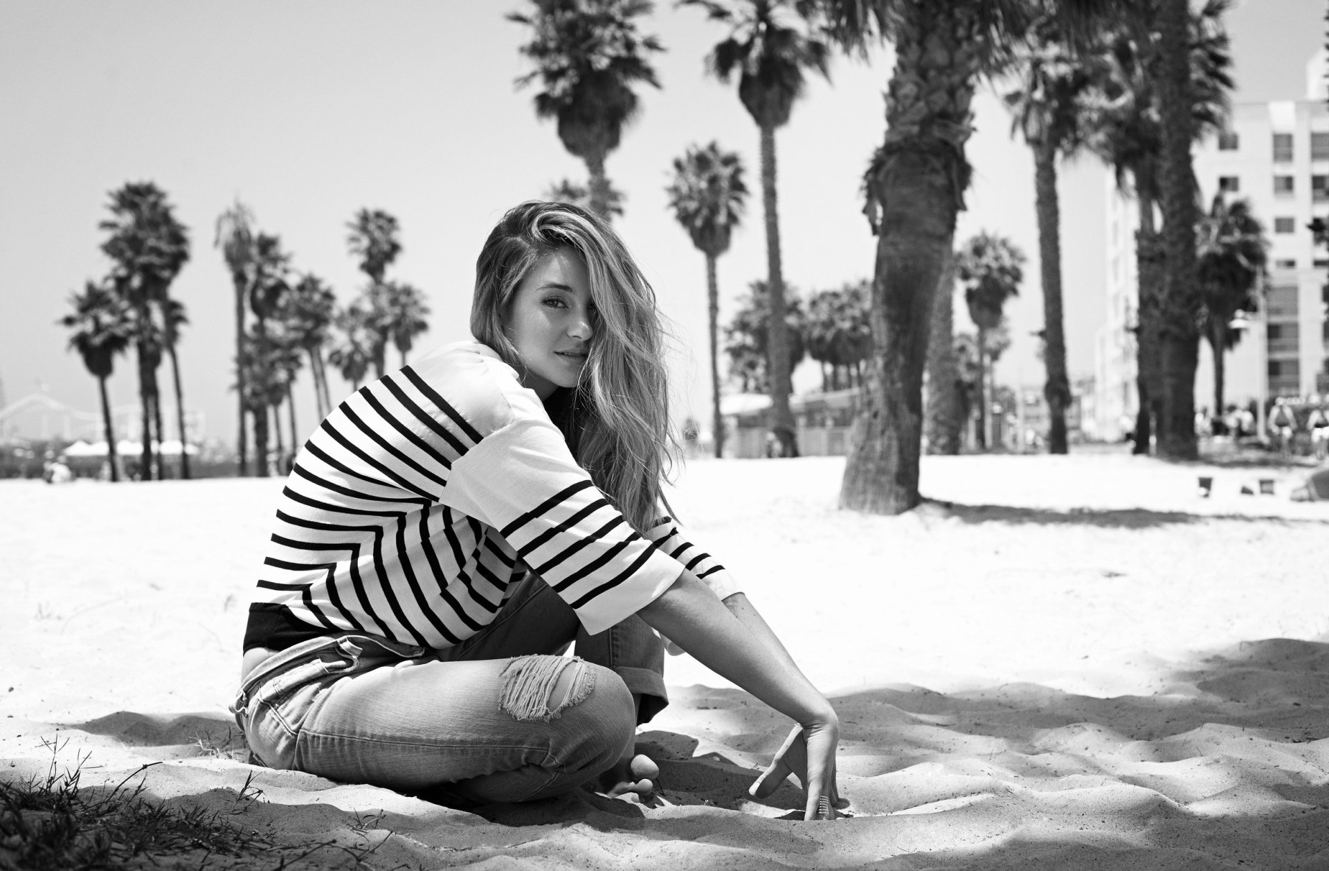 Black and white image of actress Shailene Woodley sitting on the beach, playing with sand, surrounded by palm trees, showcasing a serene moment in an American coastal setting.