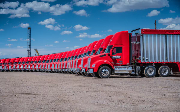 HD PC desktop wallpaper showcasing a lineup of bright red Kenworth trucks under a blue sky with clouds on a dirt lot.