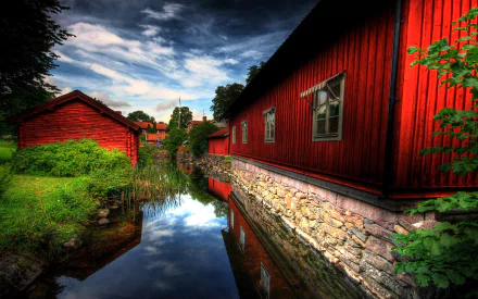 A serene village scene features red wooden houses along a reflective waterway, surrounded by lush grass, trees, and a cloudy sky, creating a peaceful, picturesque backdrop.