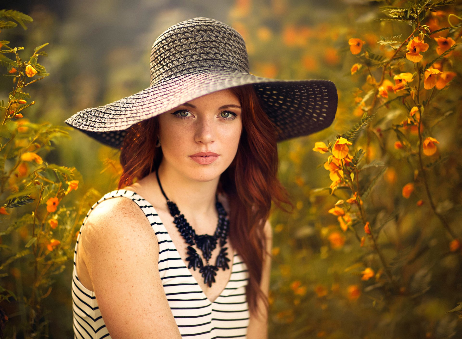A redhead woman wearing a wide-brimmed hat and a striking necklace poses amidst vibrant yellow flowers, captured in a warm and inviting setting.