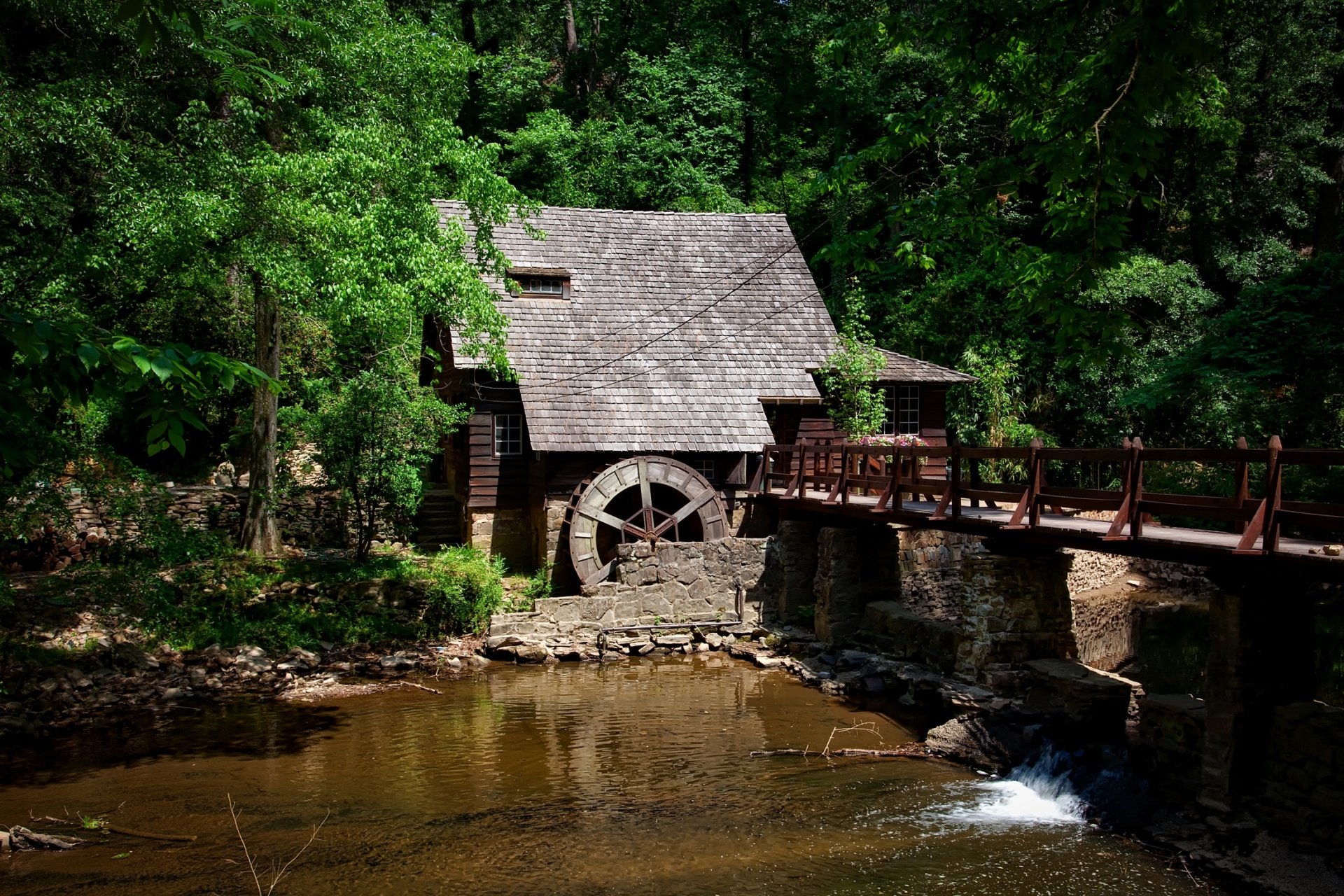 A serene forest scene featuring a watermill building beside a flowing stream, connected by a wooden bridge, captured in high-definition quality.